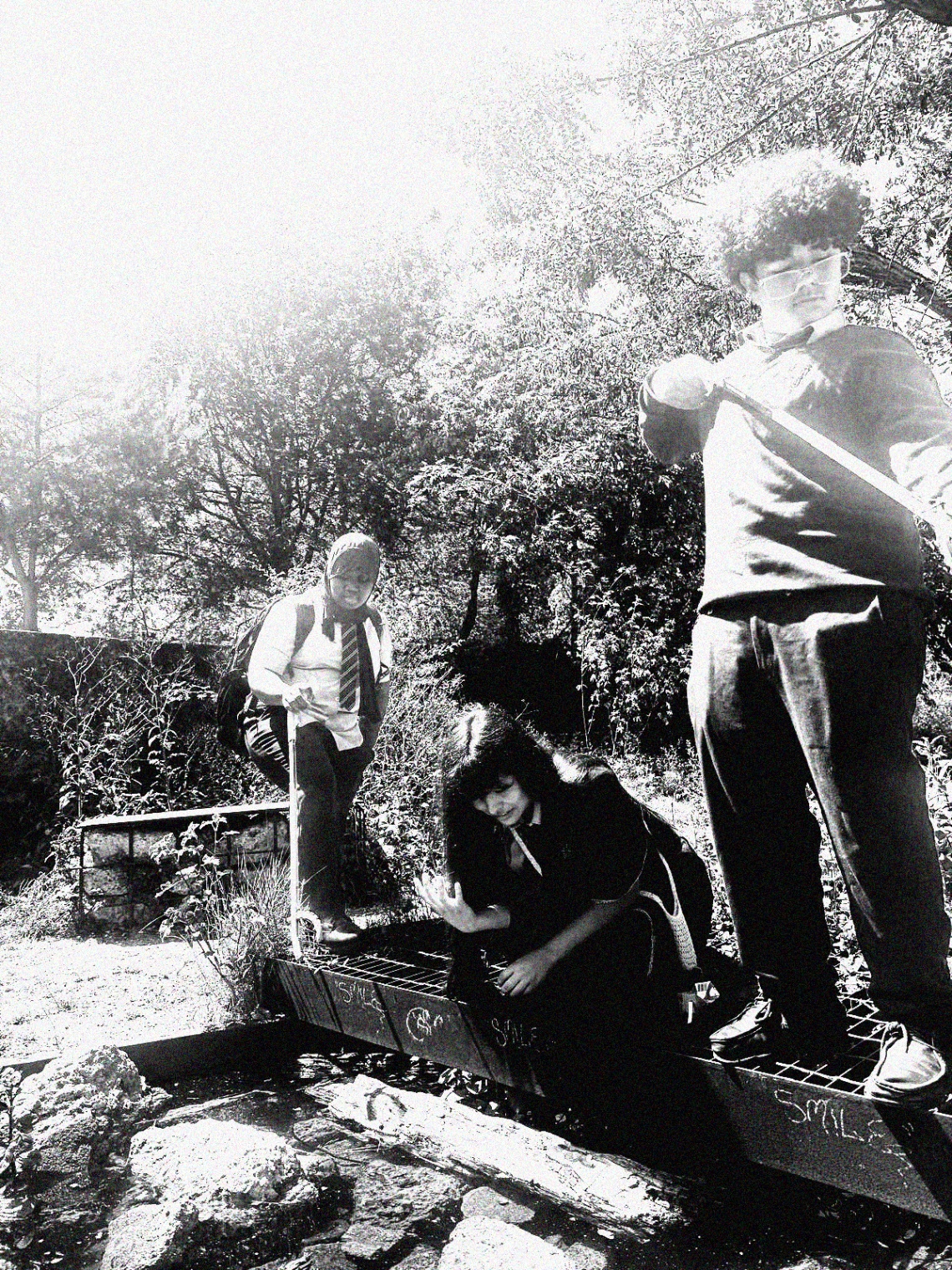 Three students on a metal grate over a small urban pond, one crouching to reach the water