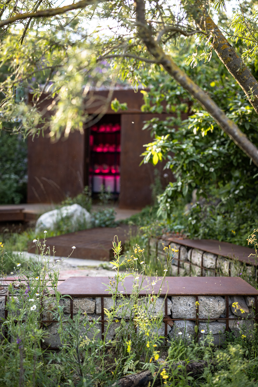 Gabion benches of reclaimed concrete framing a corten shelter glowing pink within