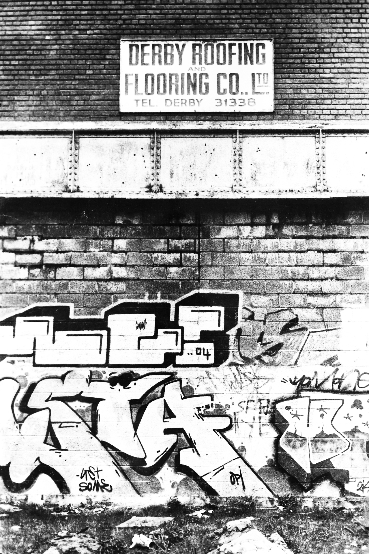 Derby brick wall and railway bridge, covered in graffiti under a hand-painted 'Derby Roofing and Flooring Co.' sign.