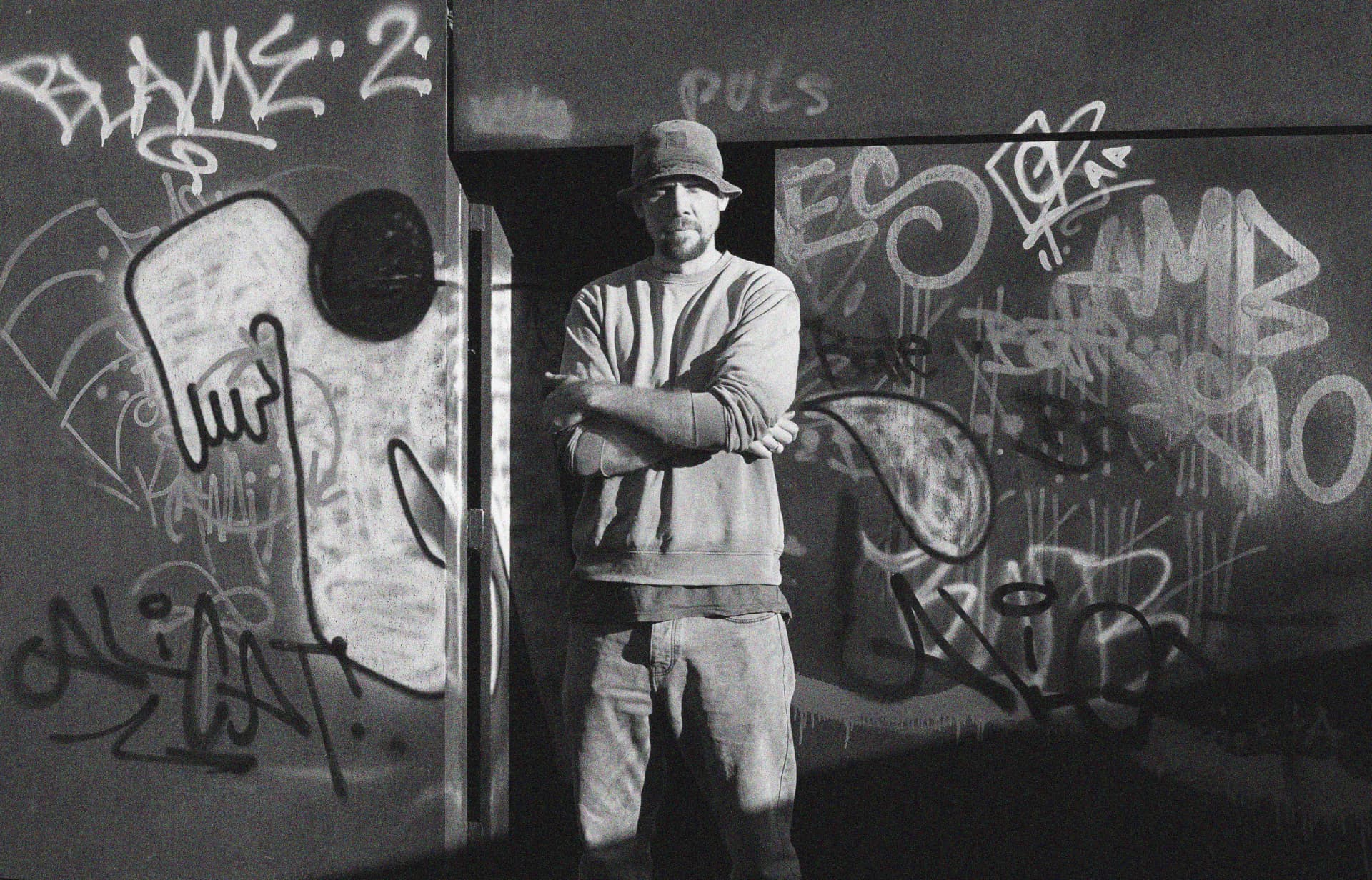 Steve Williams standing with arms folded in front of a graffiti wall, black and white