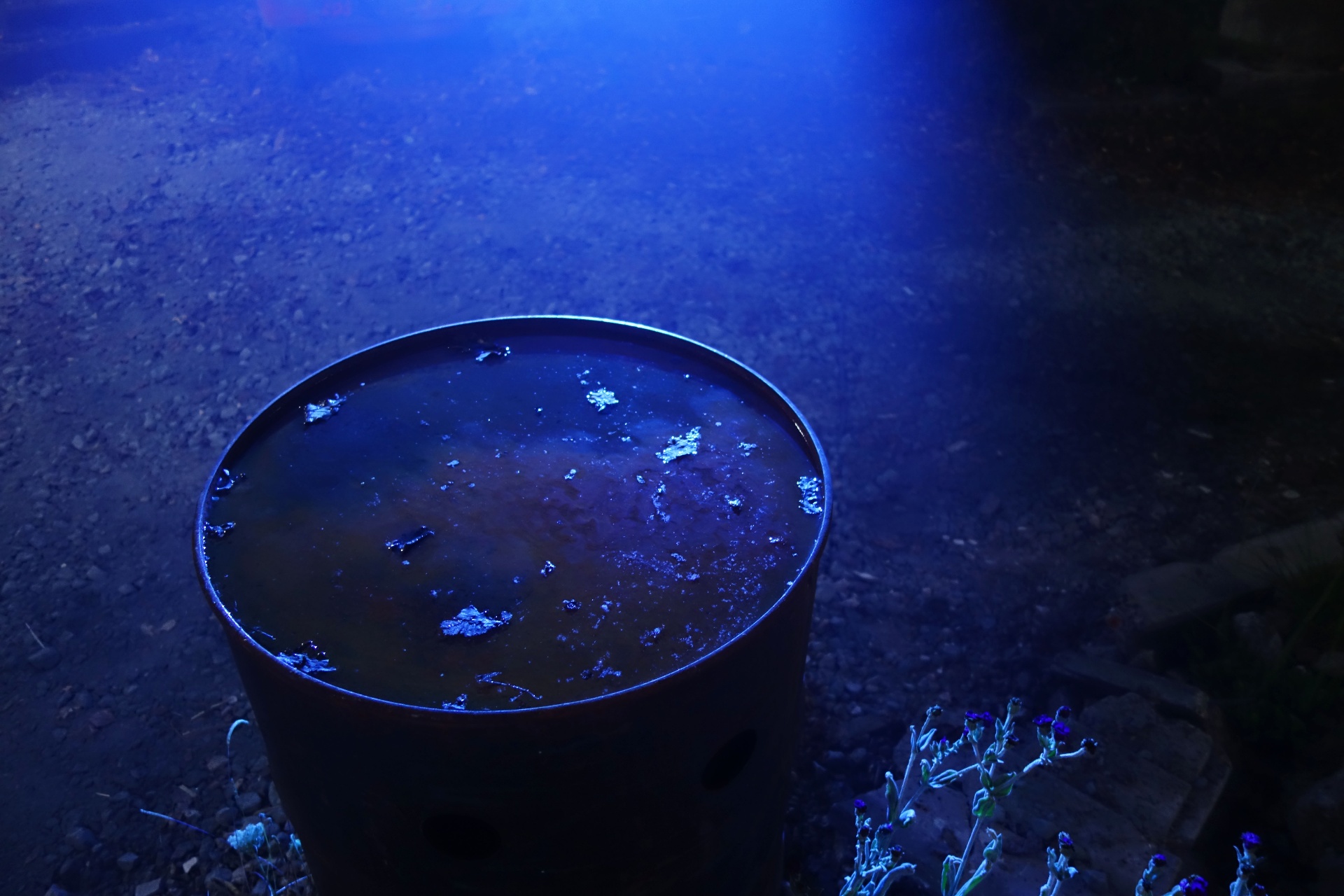 An oil drum of still water reflecting blue light on gravel