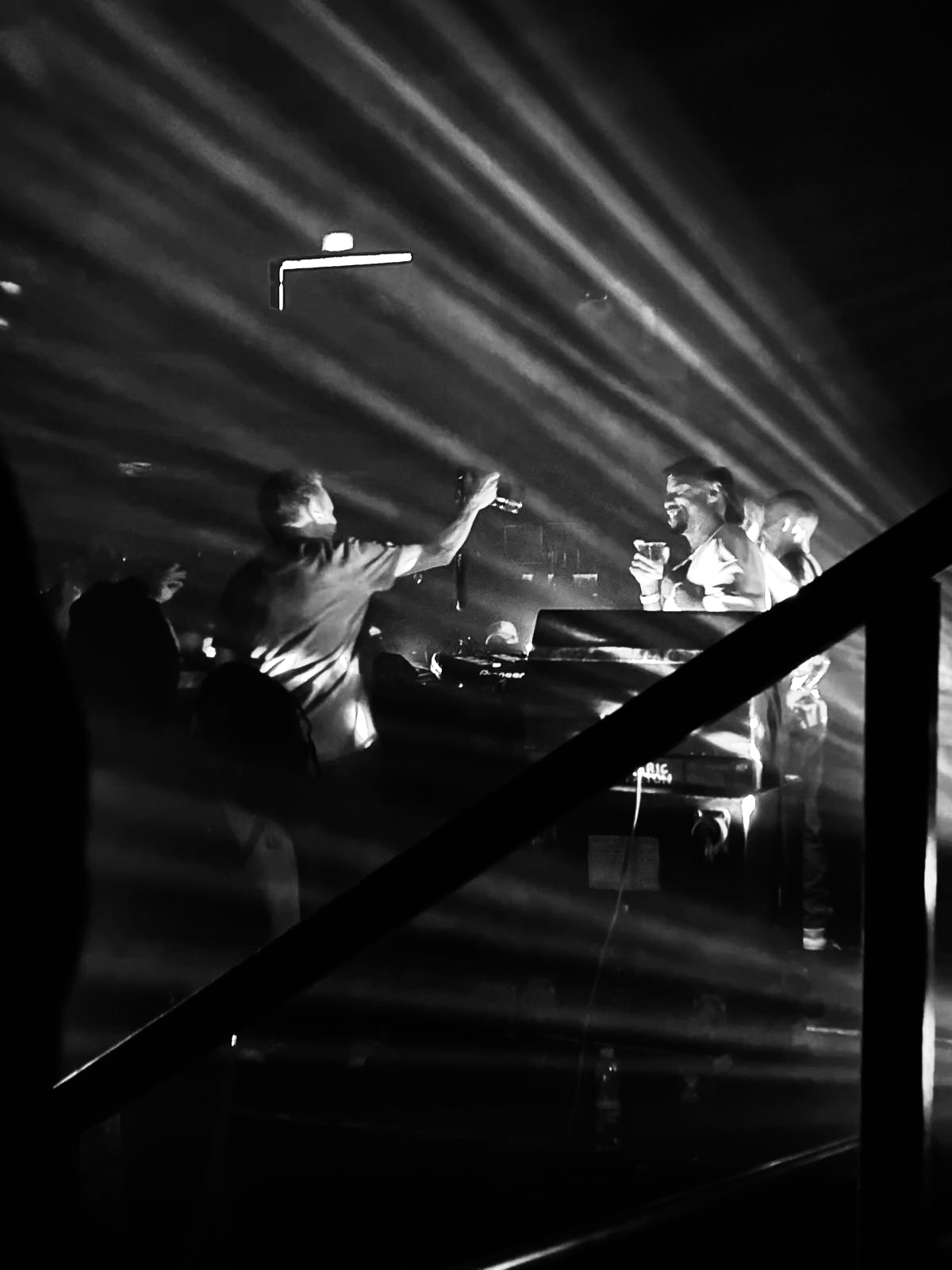 Black-and-white photograph of a DJ booth cut by beams of light, hands raised in the crowd.