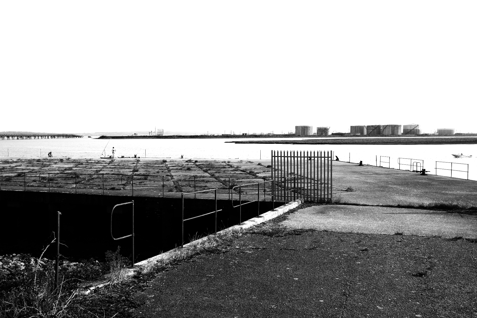 A concrete dock edge looking out across the Thames estuary to distant industrial storage tanks.