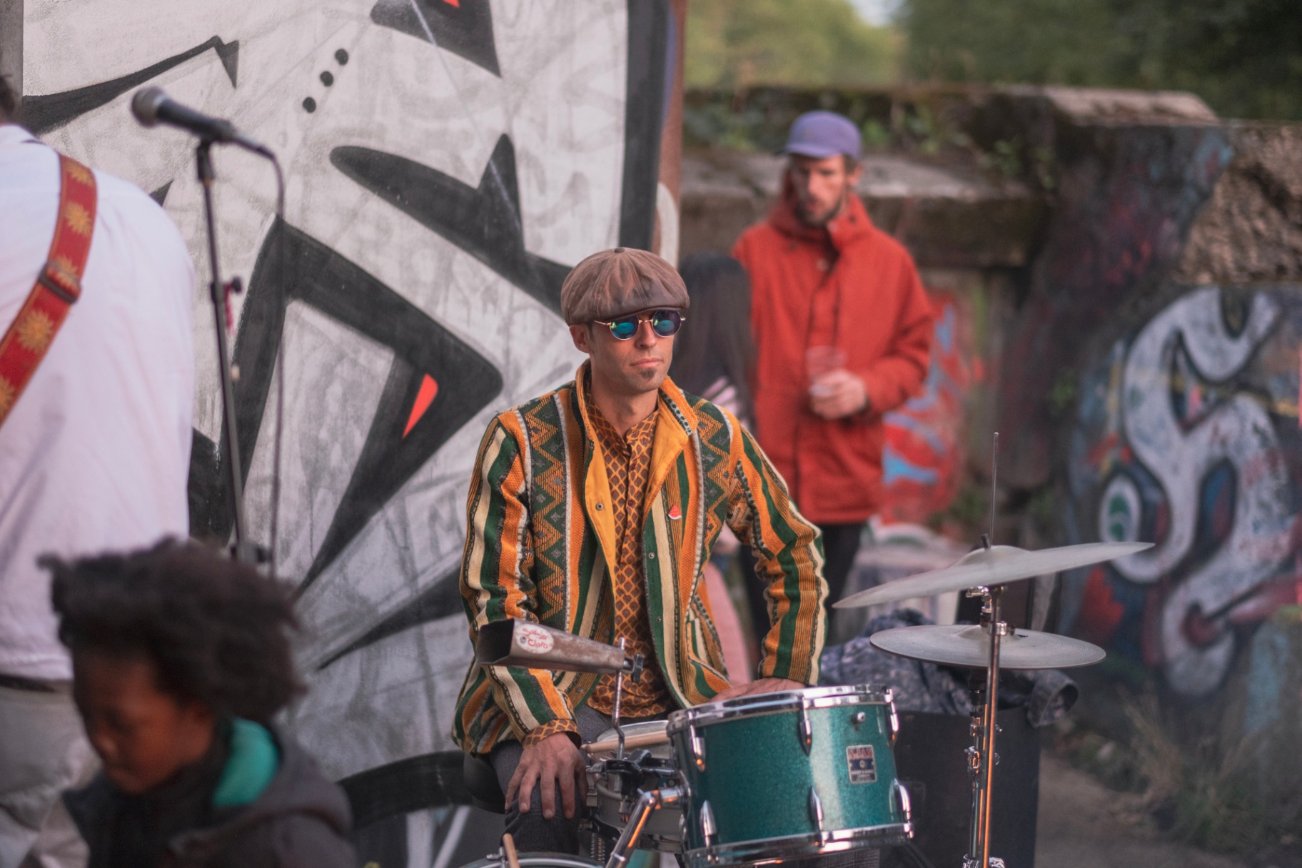 Drummer in a patterned jacket playing against a graffitied wall.